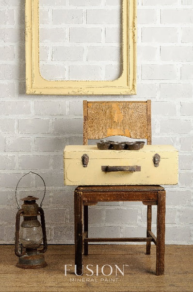 Fusion Mineral Paint | A buttermilk cream painted briefcase sitting on a wooden chair in front of a white brick wall with a picture frame painted in Buttermilk cream.